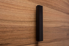L-form black cabinet handle on walnut door, emphasizing the smooth finish and geometric design.