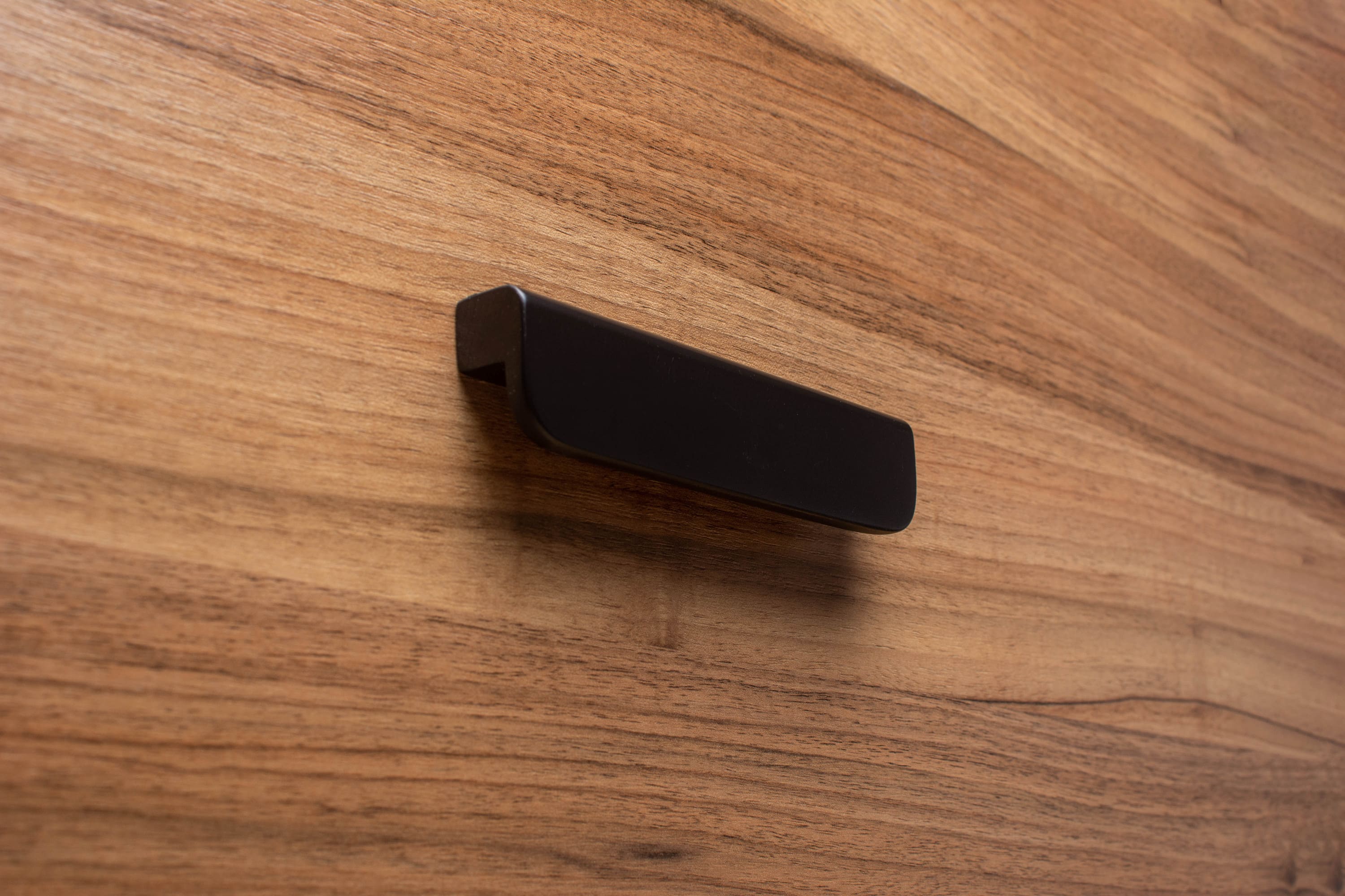 Rounded black cabinet handle with wide grip on walnut wood, offering a sleek contrast.