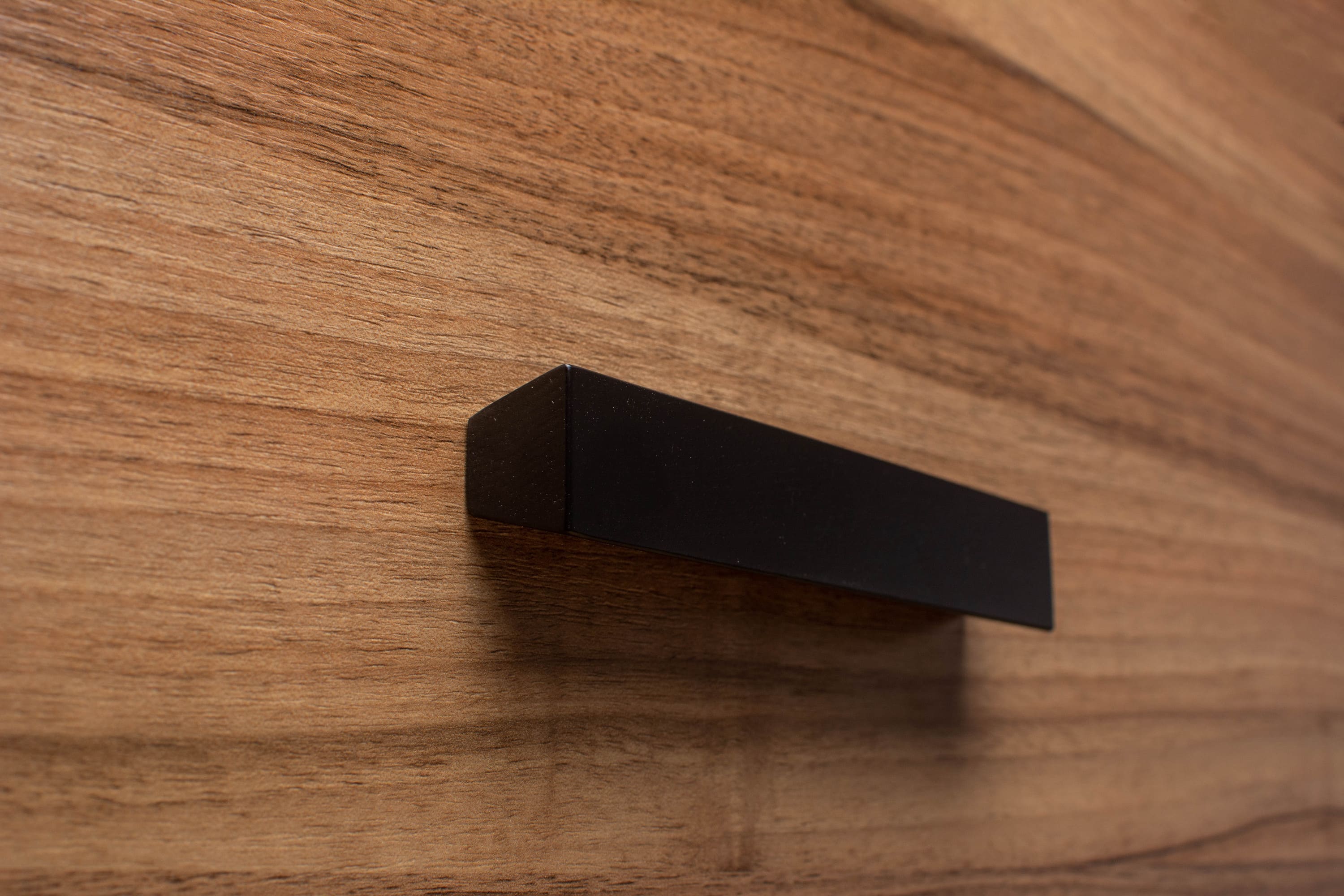 Trapezoidal black cabinet handle on walnut drawer, adding geometric flair to modern furniture design.